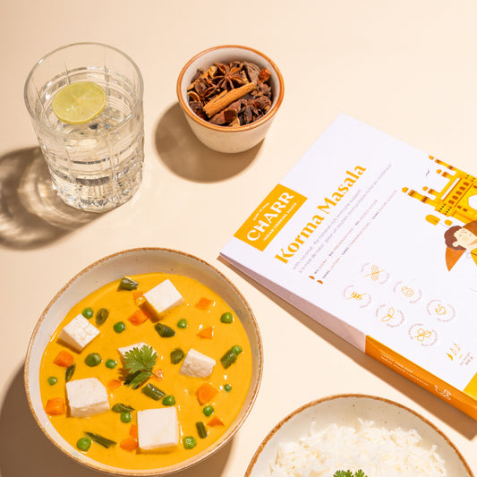 Yellow soup with tofu and vegetables, glass of water, spices, and 'Charr' cookbook on a beige surface.